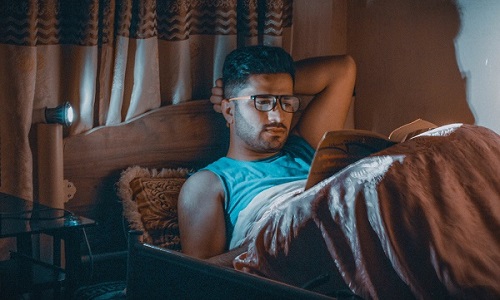 Man wearing glasses and lying down in bed and reading a book