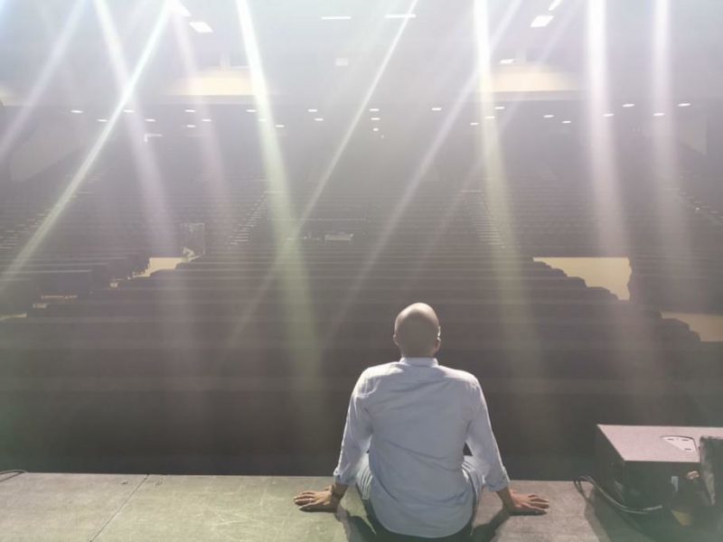 Ahmed sitting on the edge of the stage under spotlights and looking out at empty seats