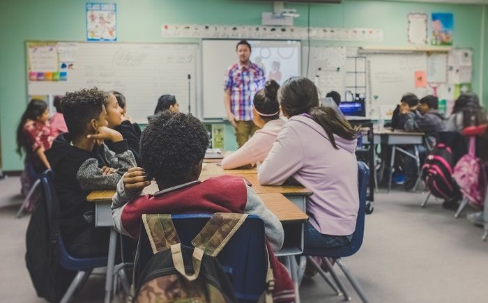 Klassenzimmer mit Kindern, die sich hinsetzen, und einem Lehrer, der aufsteht und mit ihnen spricht