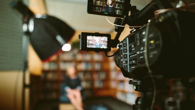 Camera pointing to a blurred person sitting in front of book case and under lamp