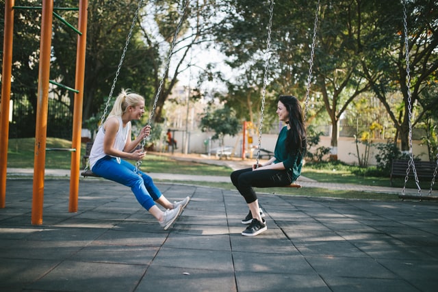 Photo of two white women in a playground sitting on swings side-by-side, facing each other and laughing during a conversation