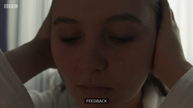 Clip of Casualty where Jade is putting her hands over her ears and hearing aids, with the caption "feedback" at the bottom