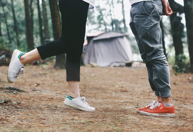 Photo of 2 people's legs, one standing on one leg, the other standing opposite on two feet, with a camp ground in the background